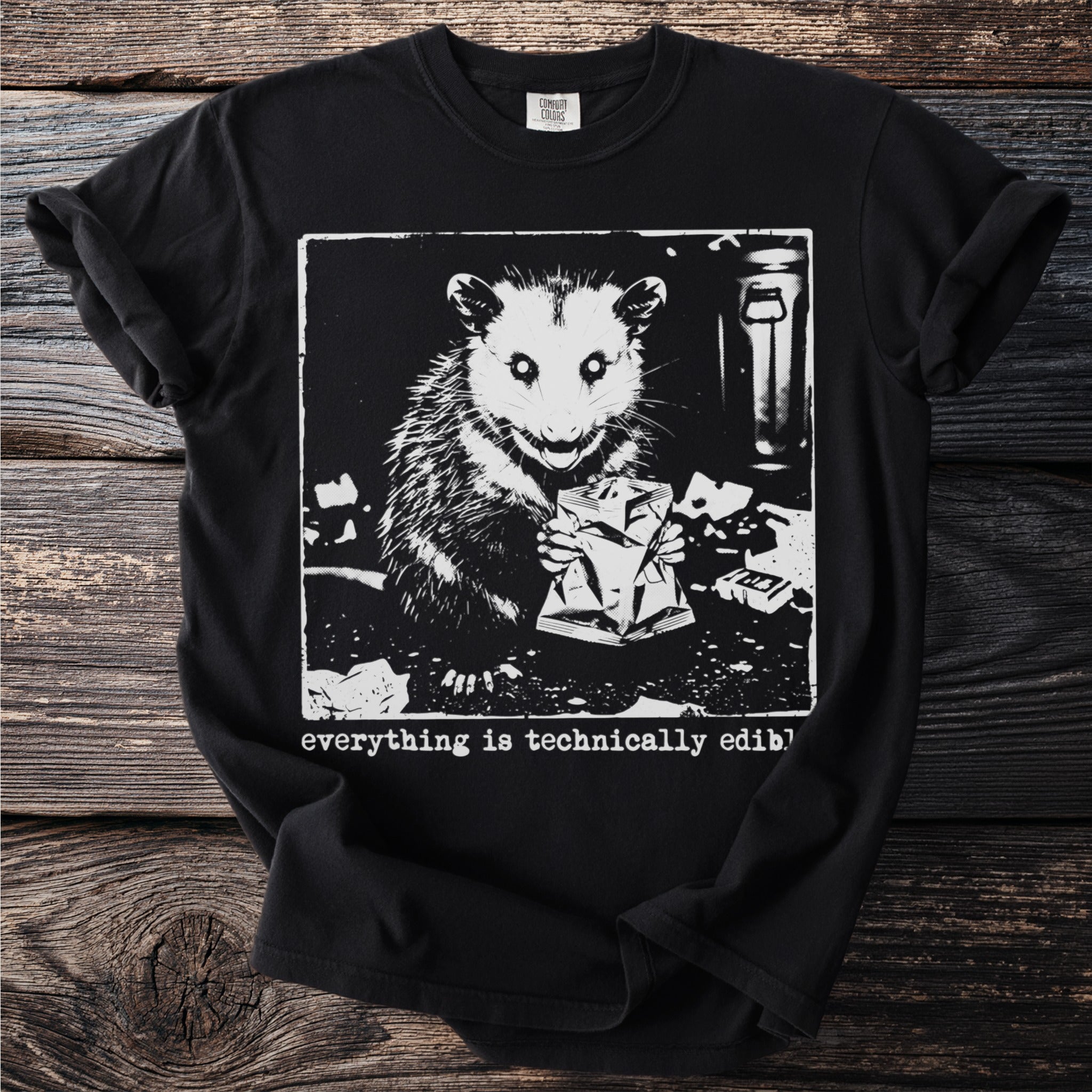 Black t-shirt with a print of a possum holding a bag of chips with text "everything is technically edib!".