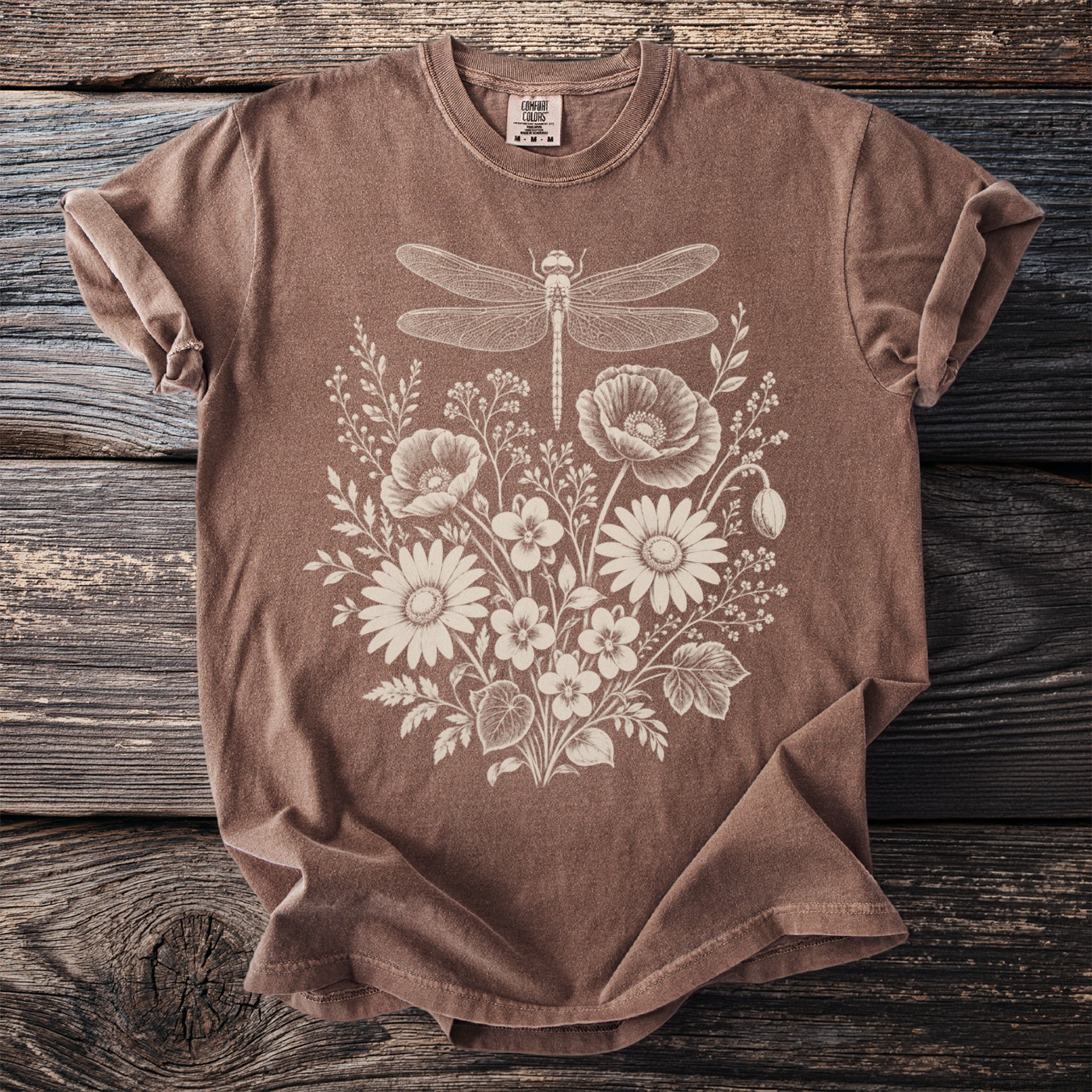 Brown t-shirt with a white dragonfly and floral print on a wooden background.