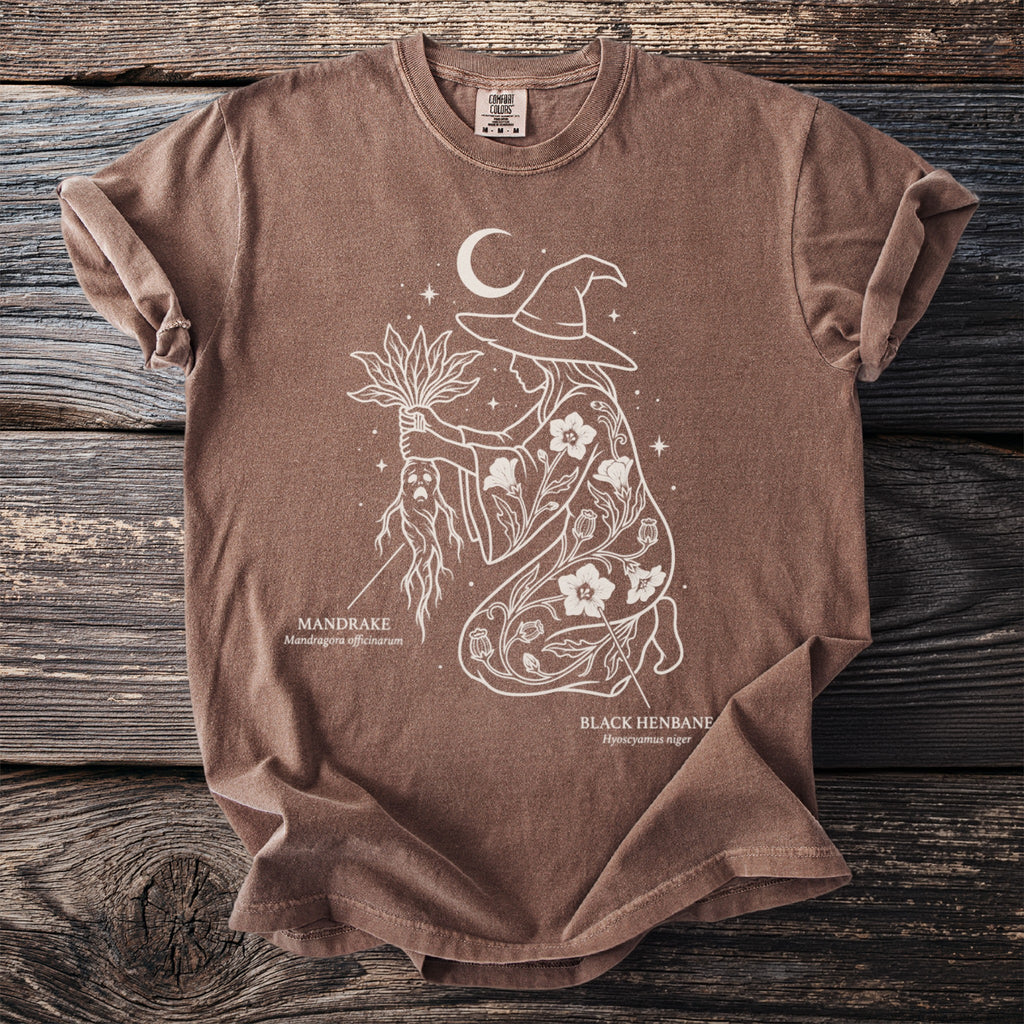 Brown t-shirt with a white line drawing of a witch holding mandrake and black henbane.