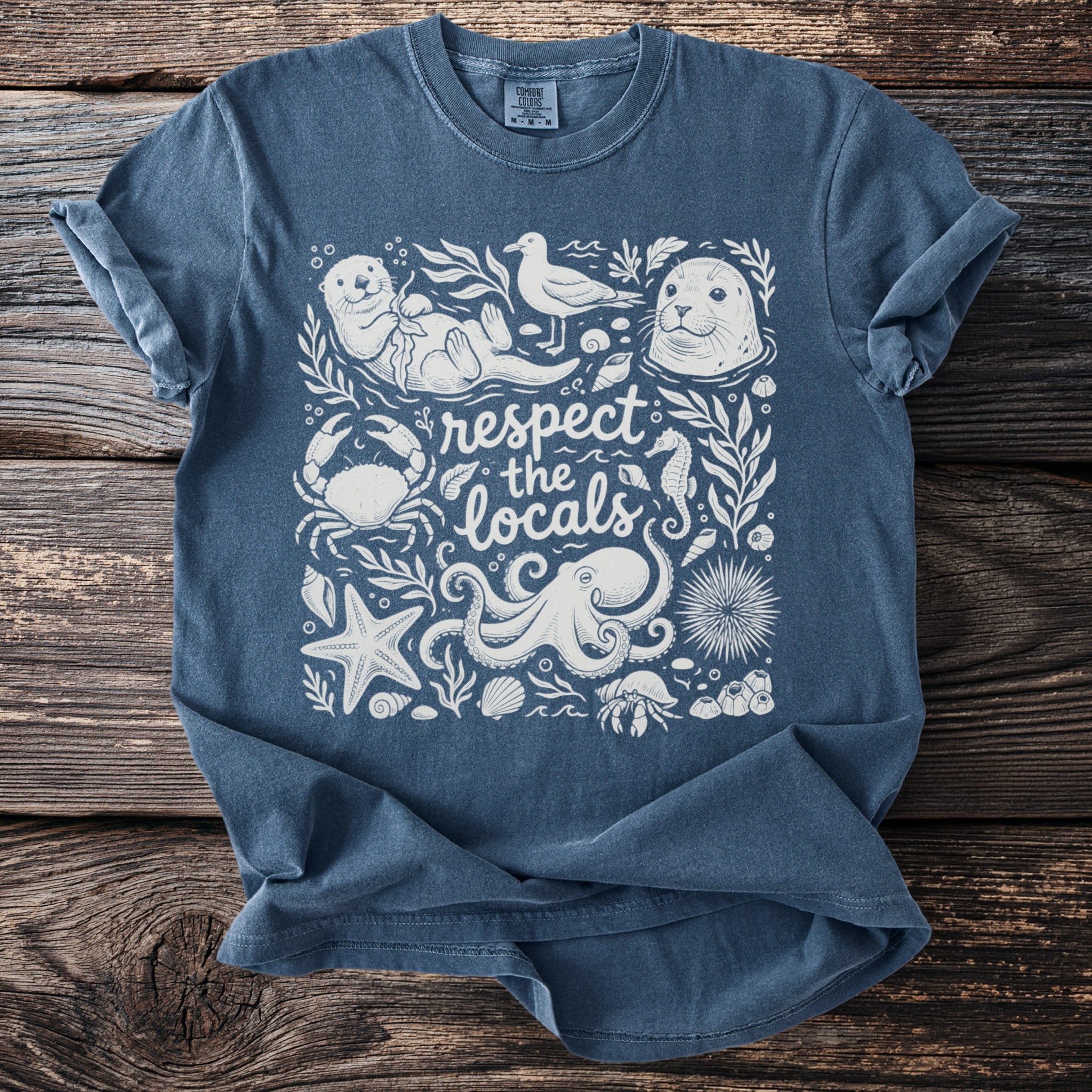 Blue t-shirt with white ocean themed art including a sea otter, seal, and "respect the locals" text.