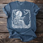 Blue t-shirt with white illustration of woman spinning yarn under a full moon.