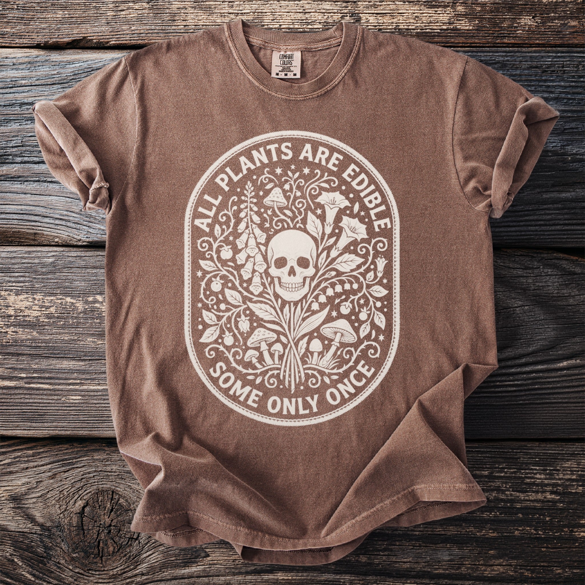 Brown t-shirt with white ornamental design of skull, plants, and mushrooms with text.