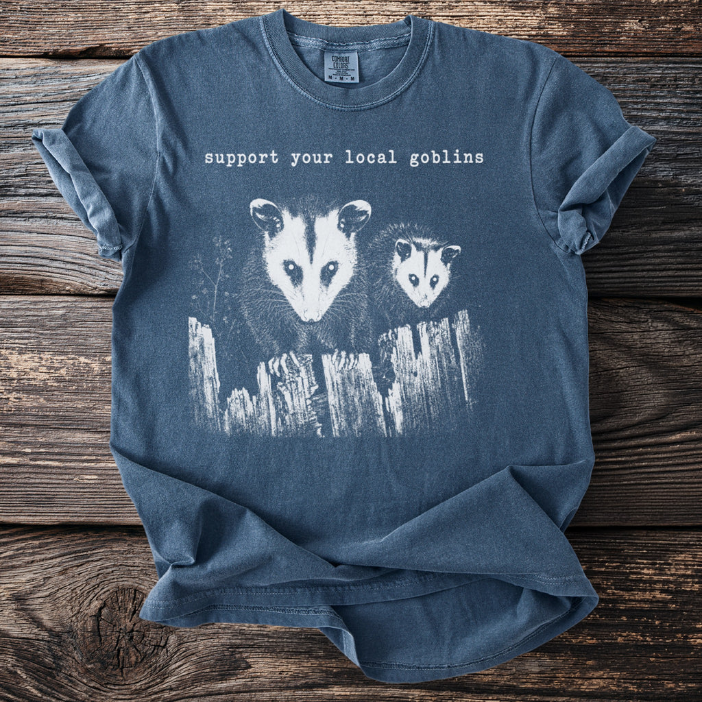 Blue t-shirt with "Support your local goblins" text and two opossums behind a fence.