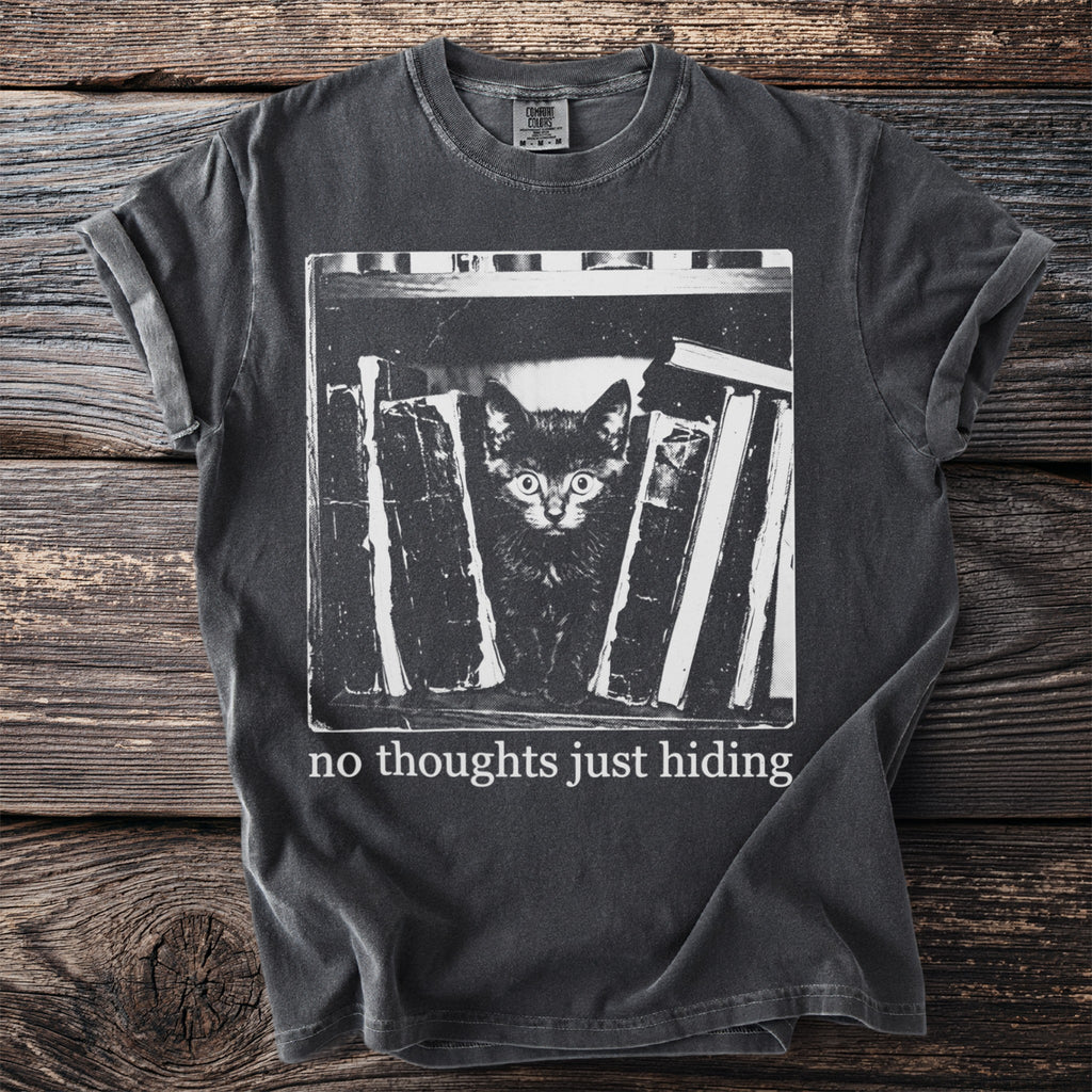 Black t-shirt with a screen print of a kitten peeking from behind books and text, "no thoughts just hiding."
