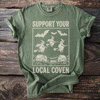 Green t-shirt with "Support Your Local Coven" text and witches flying over houses.