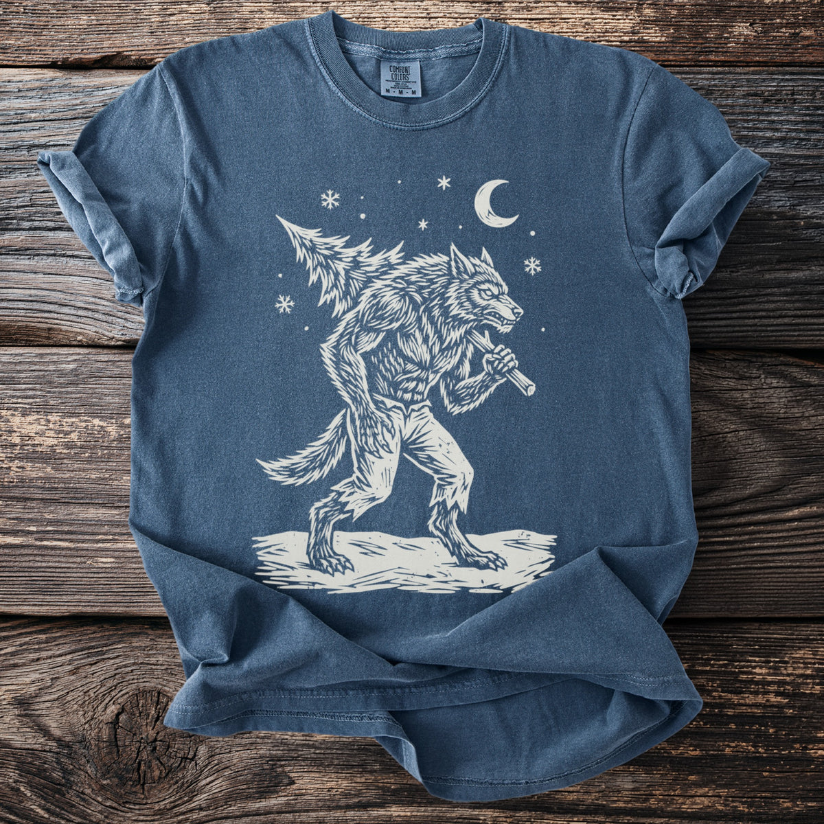 Denim blue t-shirt with a cream folk-cut print: a snarling werewolf in torn trousers walks through the snow with a fir tree over his shoulder.