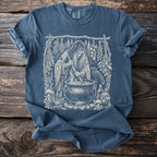 Witch stirring a cauldron with herbs, raven, and potion bottles illustration on t-shirt.