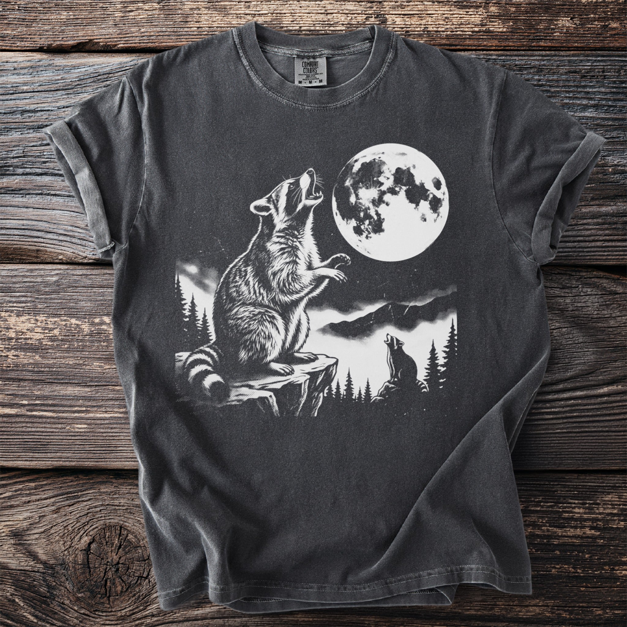 Graphic t-shirt with raccoon howling at full moon in mountainous forest scene.