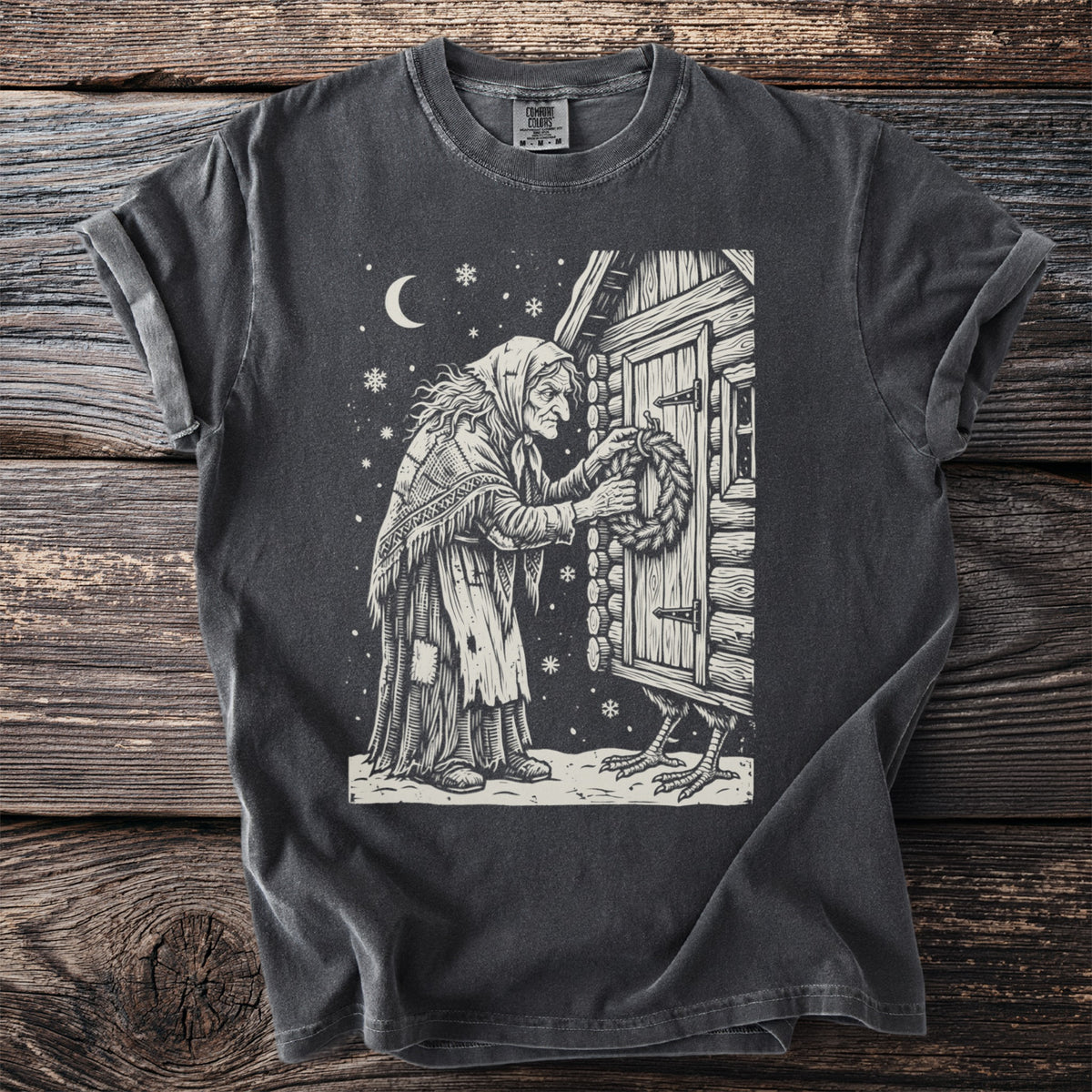 Pepper gray t-shirt with a cream folk-cut print: Baba Yaga hangs a wreath on the door of her log hut, one chicken leg visible beneath it.