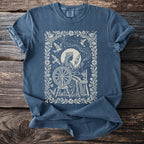 Blue t-shirt with a vintage queen spinning yarn at a wheel, surrounded by birds and constellations.