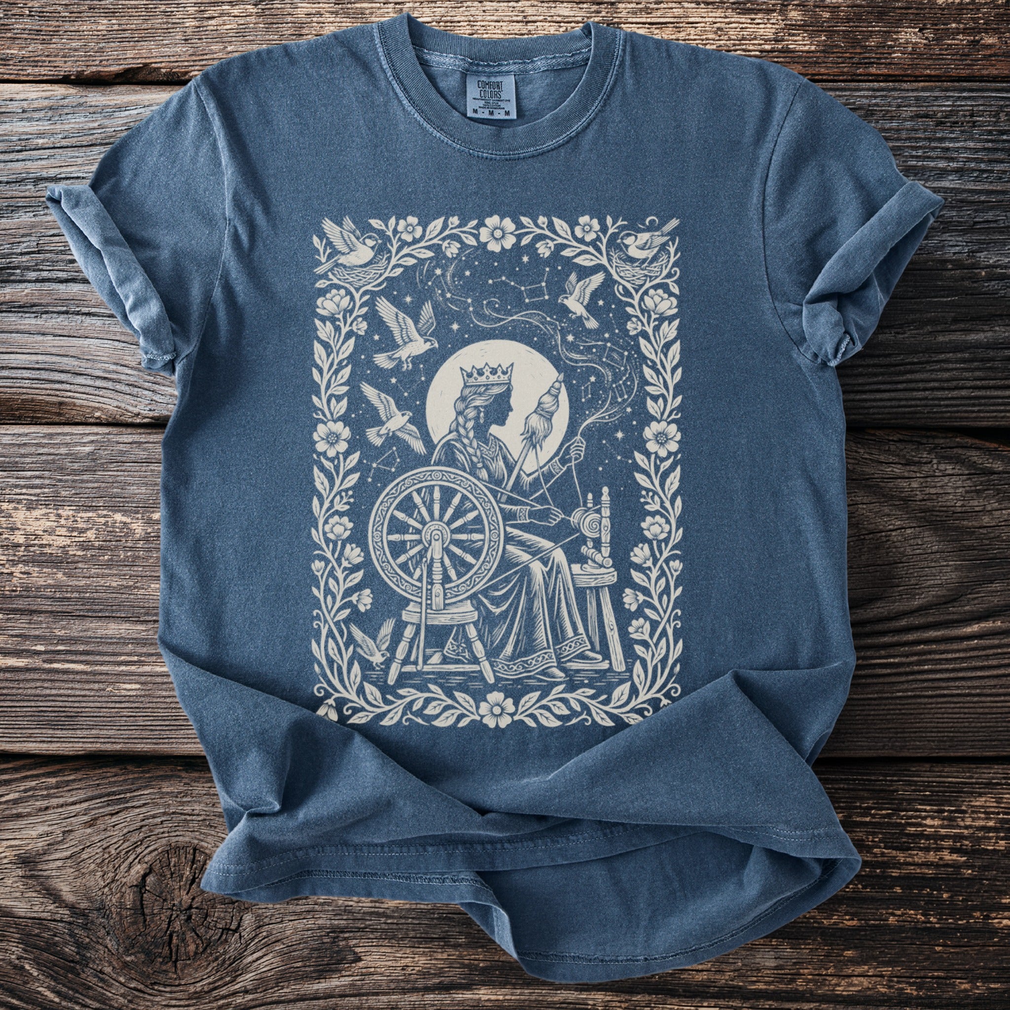 Blue t-shirt with a vintage queen spinning yarn at a wheel, surrounded by birds and constellations.