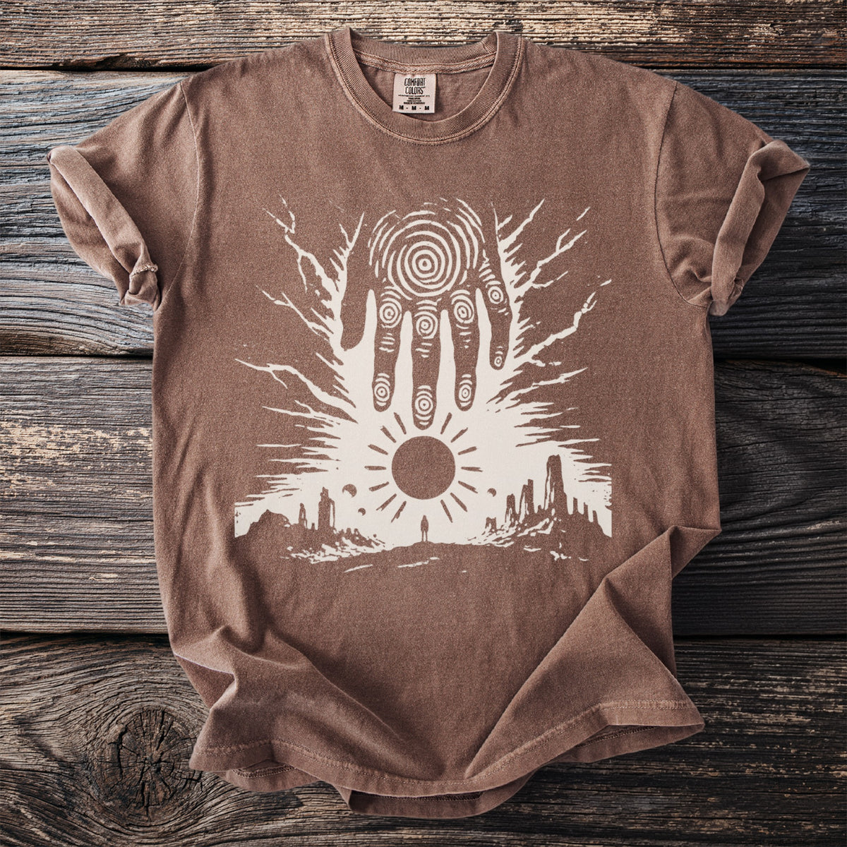Brown t-shirt with a white graphic of a hand over a desert landscape and sun.