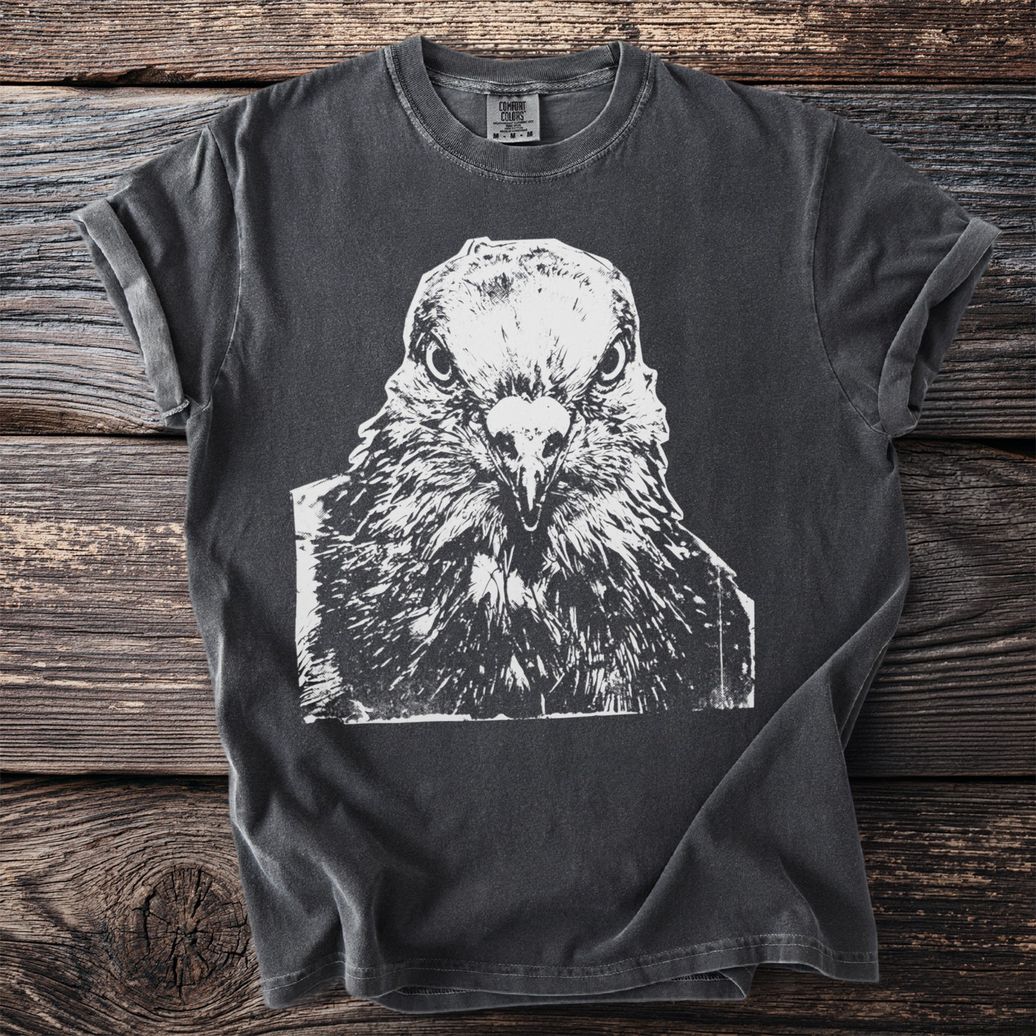 Dark t-shirt with a white close-up graphic of a pigeon's face staring straight ahead, on a wooden background.