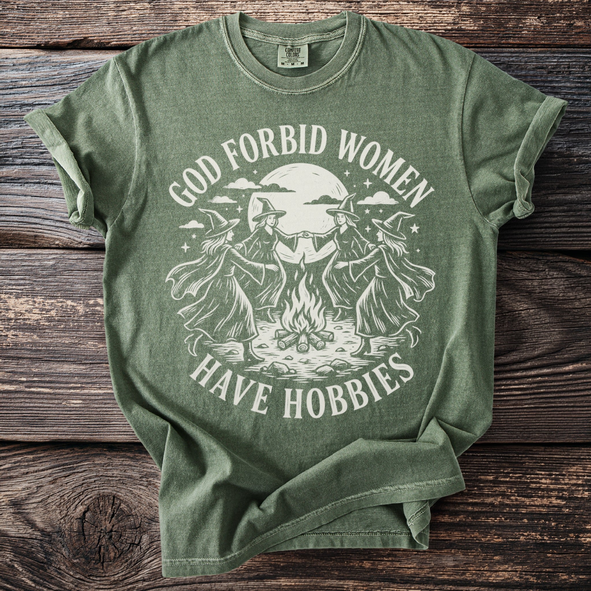 Green t-shirt with white graphic of witches dancing around a campfire under a moon.