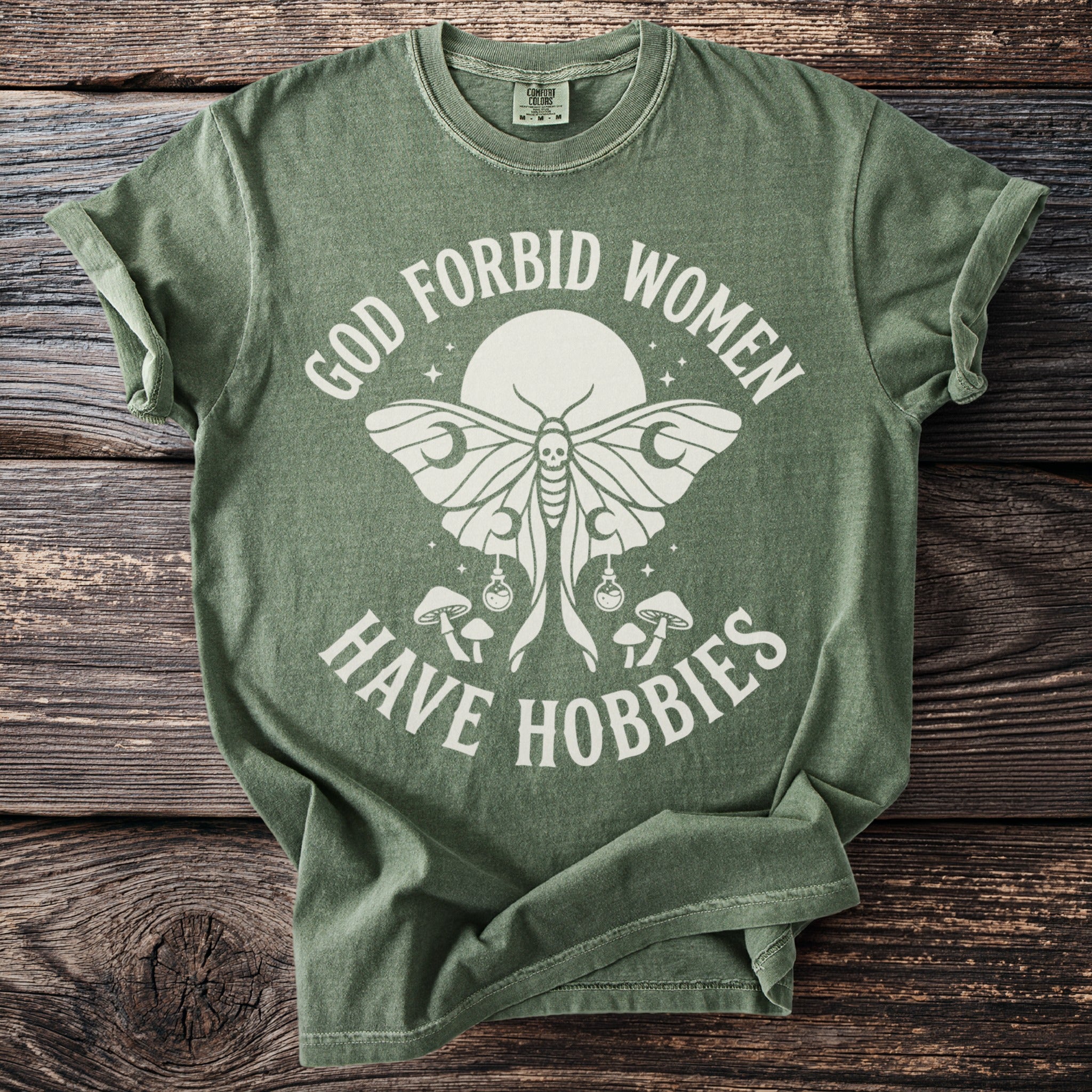Green t-shirt with a moth, mushrooms, and text "GOD FORBID WOMEN HAVE HOBBIES".