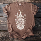 Brown t-shirt with white witch and plant graphic on an open book.