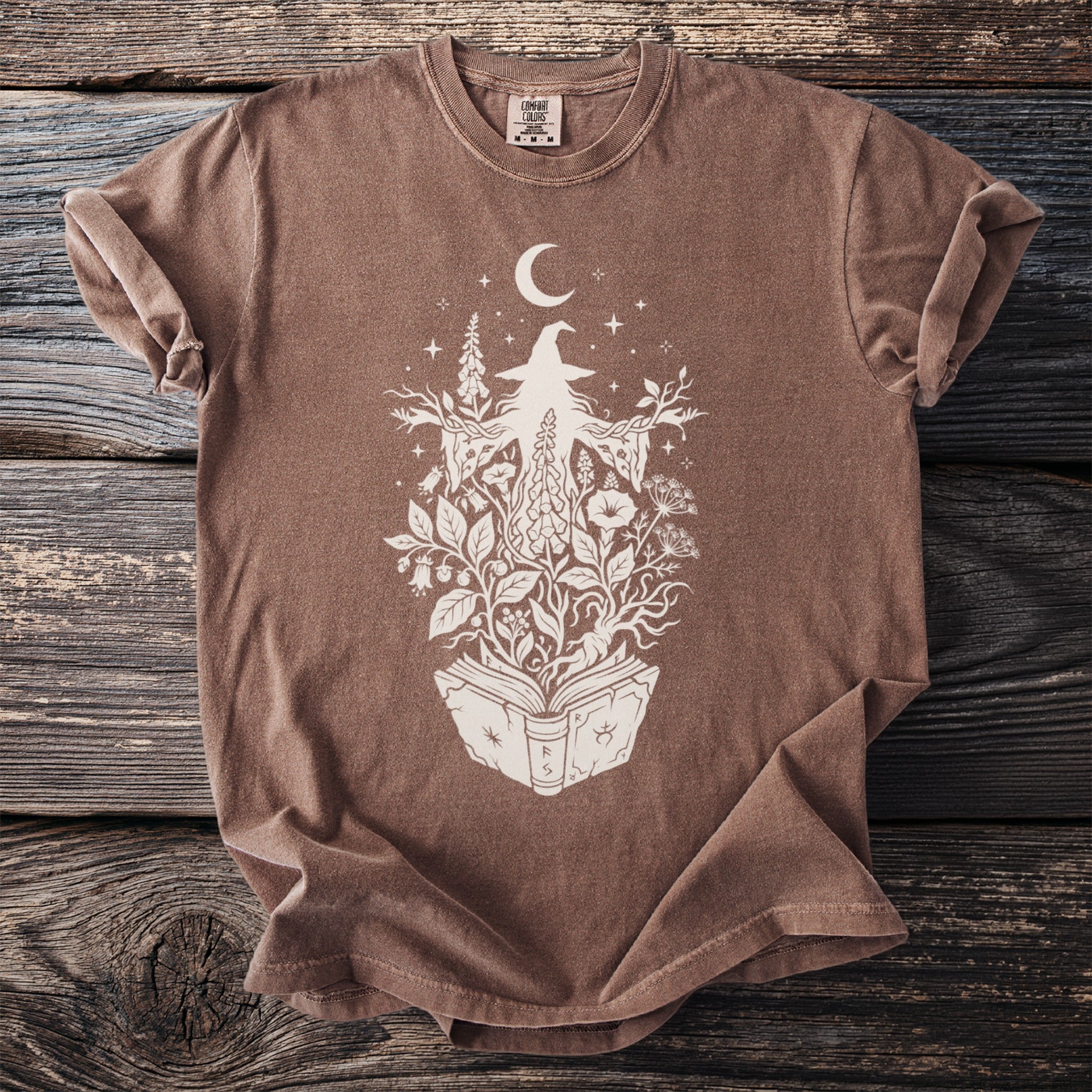 Brown t-shirt with white witch and plant graphic on an open book.