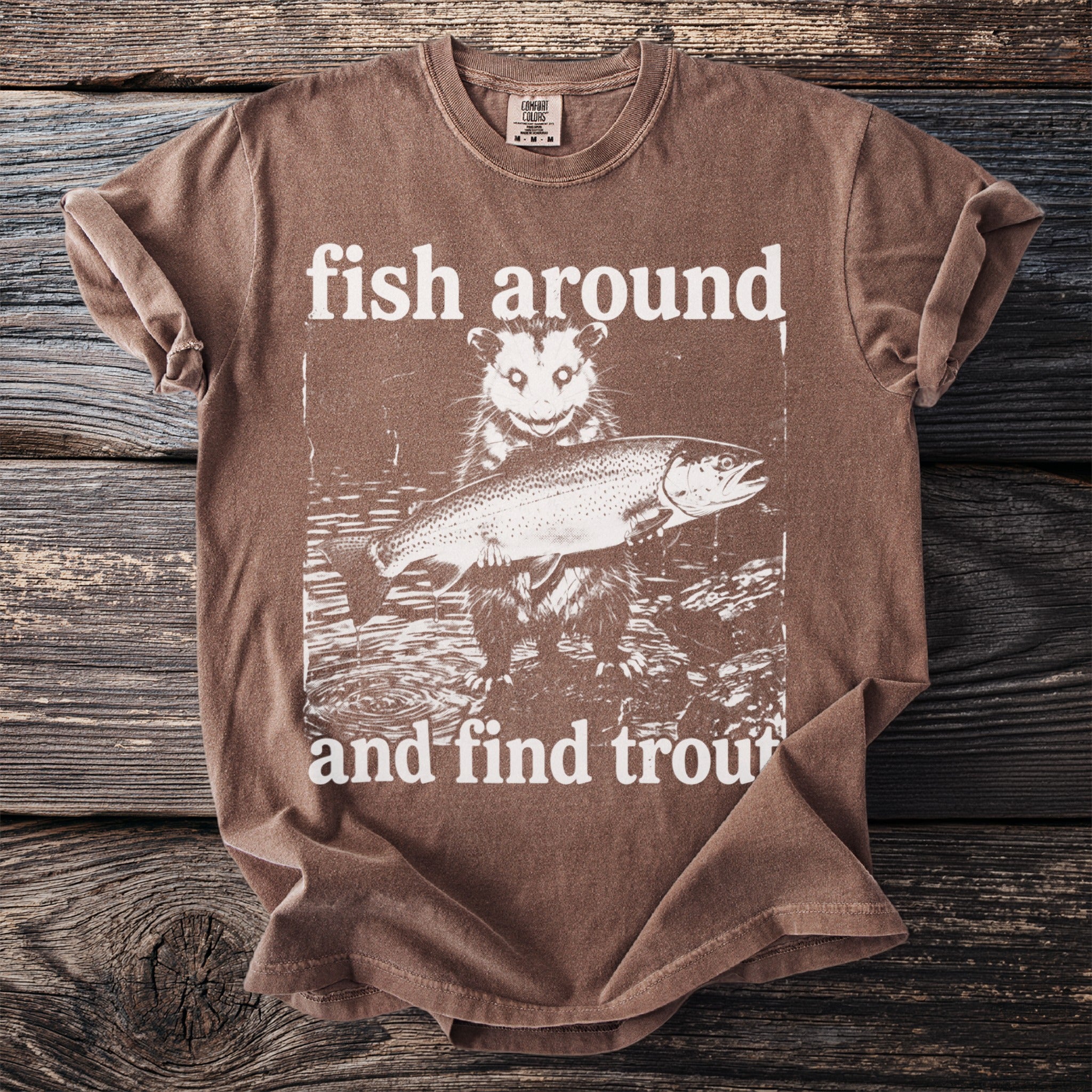 Brown t-shirt with a graphic of an opossum holding a trout and text that reads "fish around and find trout".