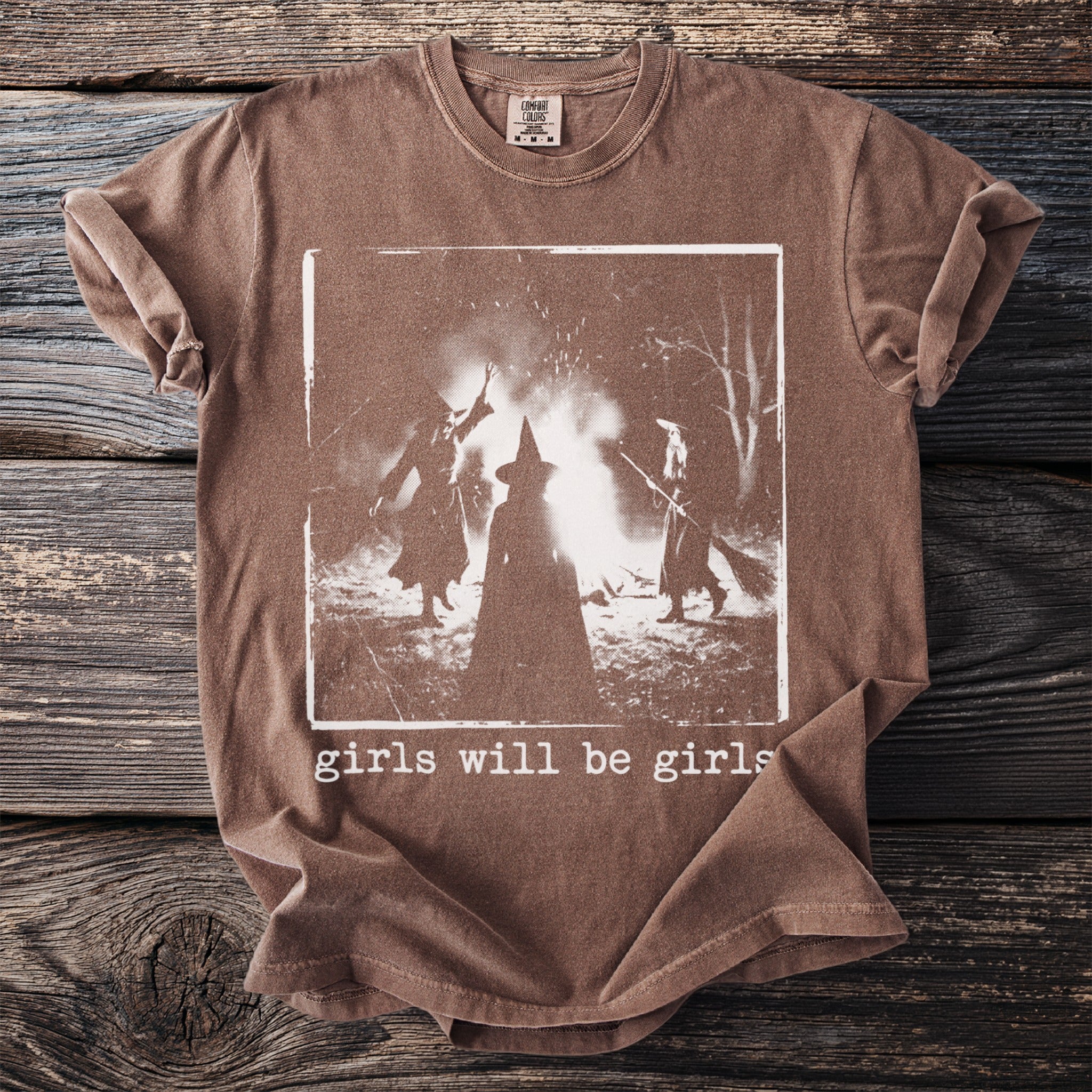 Brown t-shirt with a white graphic of three witches around a bonfire, text reads "girls will be girls".