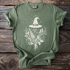 Green t-shirt with white witch hat and botanical graphic.