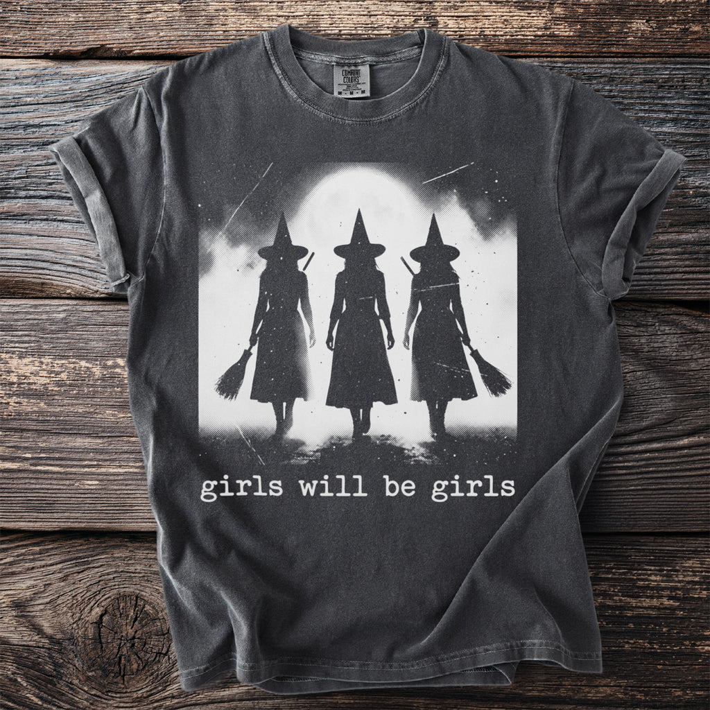 Dark t-shirt with black silhouette of three witches holding brooms under a moon with text "girls will be girls".