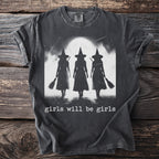 Dark t-shirt with black silhouette of three witches holding brooms under a moon with text "girls will be girls".