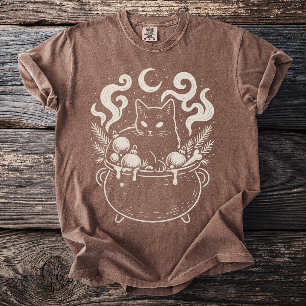 Brown t-shirt with a cream line drawing of a cat in a cauldron with ornaments.