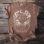 Brown t-shirt with a cream line drawing of a cat in a cauldron with ornaments.