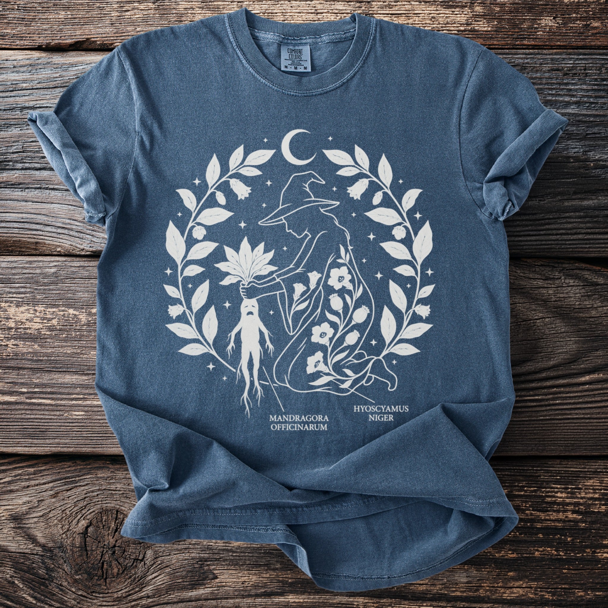 T-shirt with white line art of a witch holding a mandragora and hyoscyamus plants.