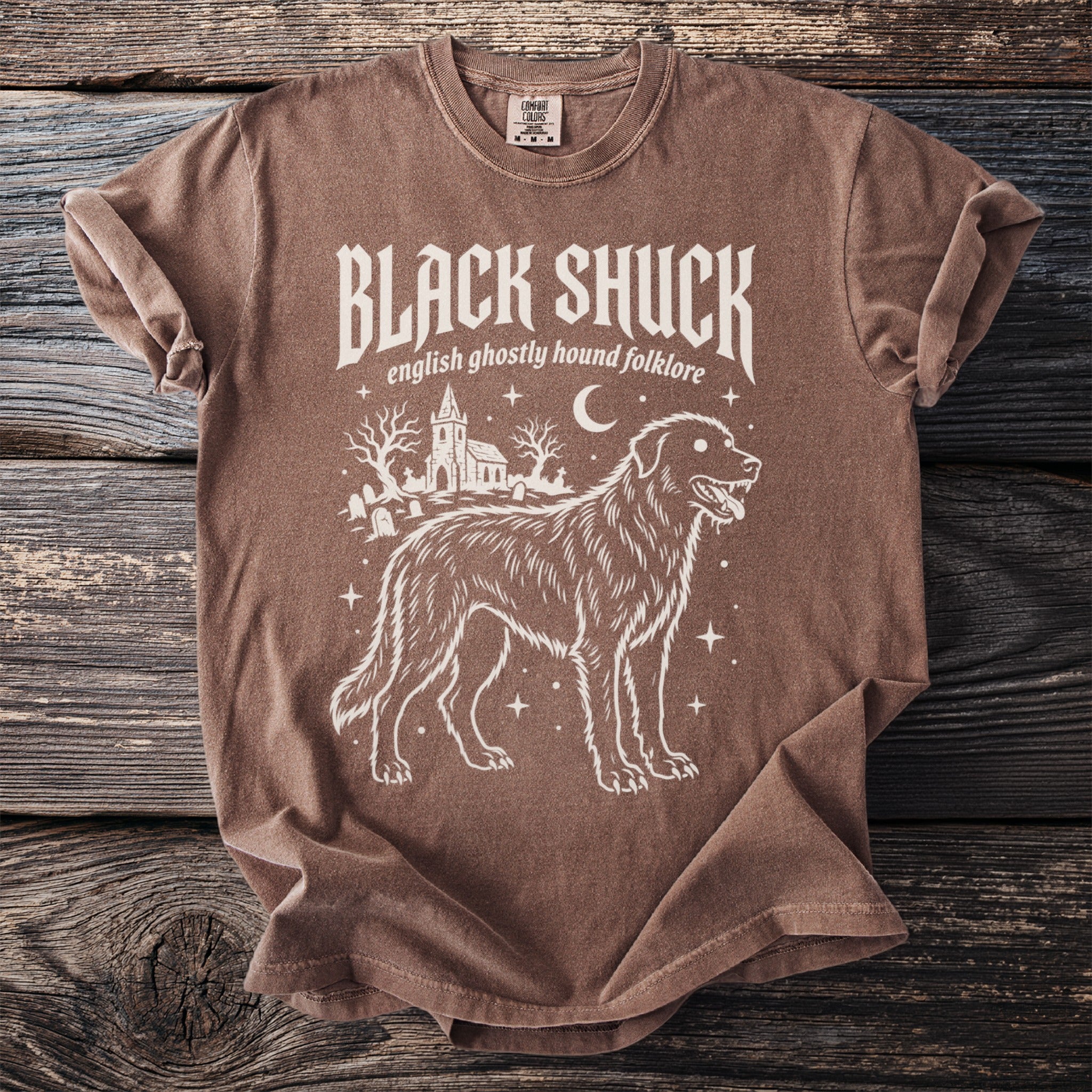Brown t-shirt with white graphic of a dog, church, and gravestones. Text reads "BLACK SHUCK english ghostly hound folklore."
