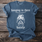 Blue t-shirt with a bat hanging upside down and text "hanging in there barely."