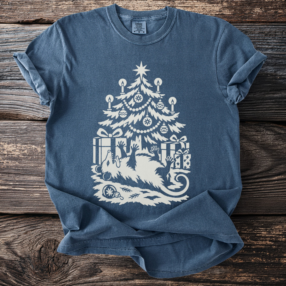 Denim blue t-shirt with a cream folk-cut print: an opossum lies on its back under a candlelit Christmas tree, playing dead among the presents.