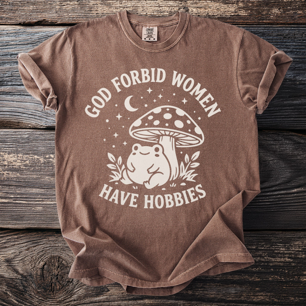 Brown t-shirt with white text and graphic of frog and mushroom saying "God forbid women have hobbies".
