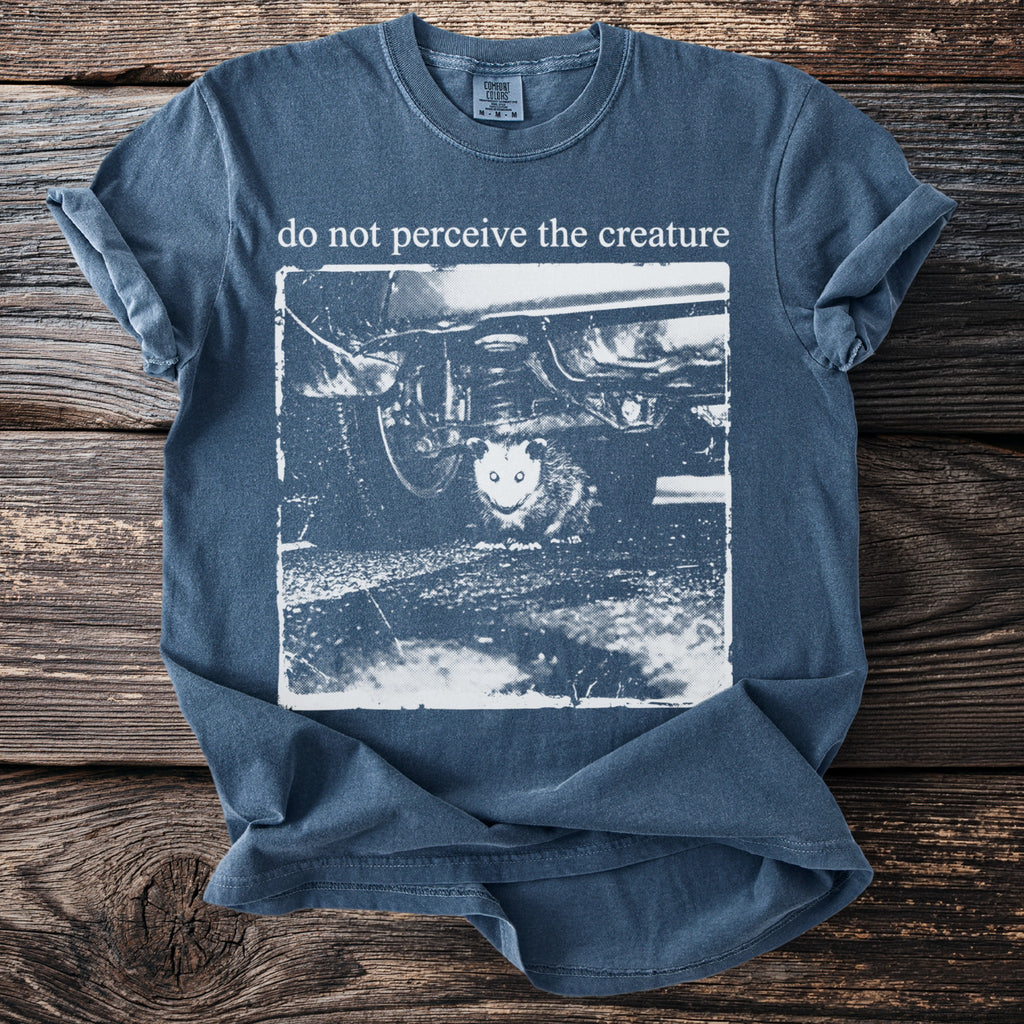 Blue shirt with a graphic of an opossum under a car and text "do not perceive the creature."