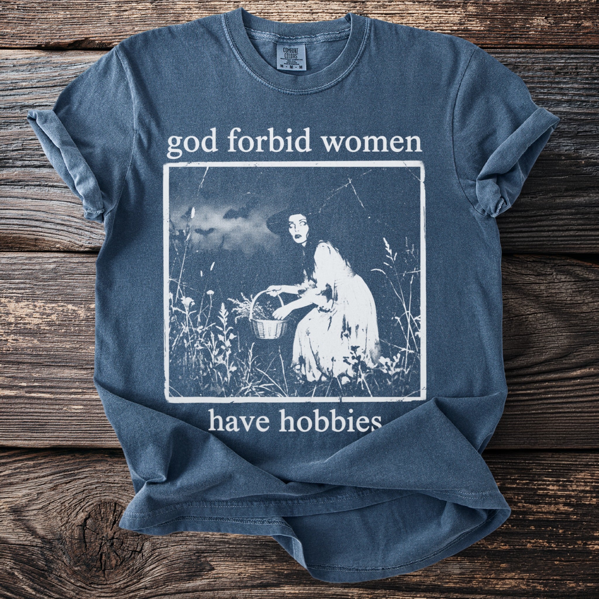 Blue t-shirt with text "god forbid women have hobbies" and a vintage illustration.