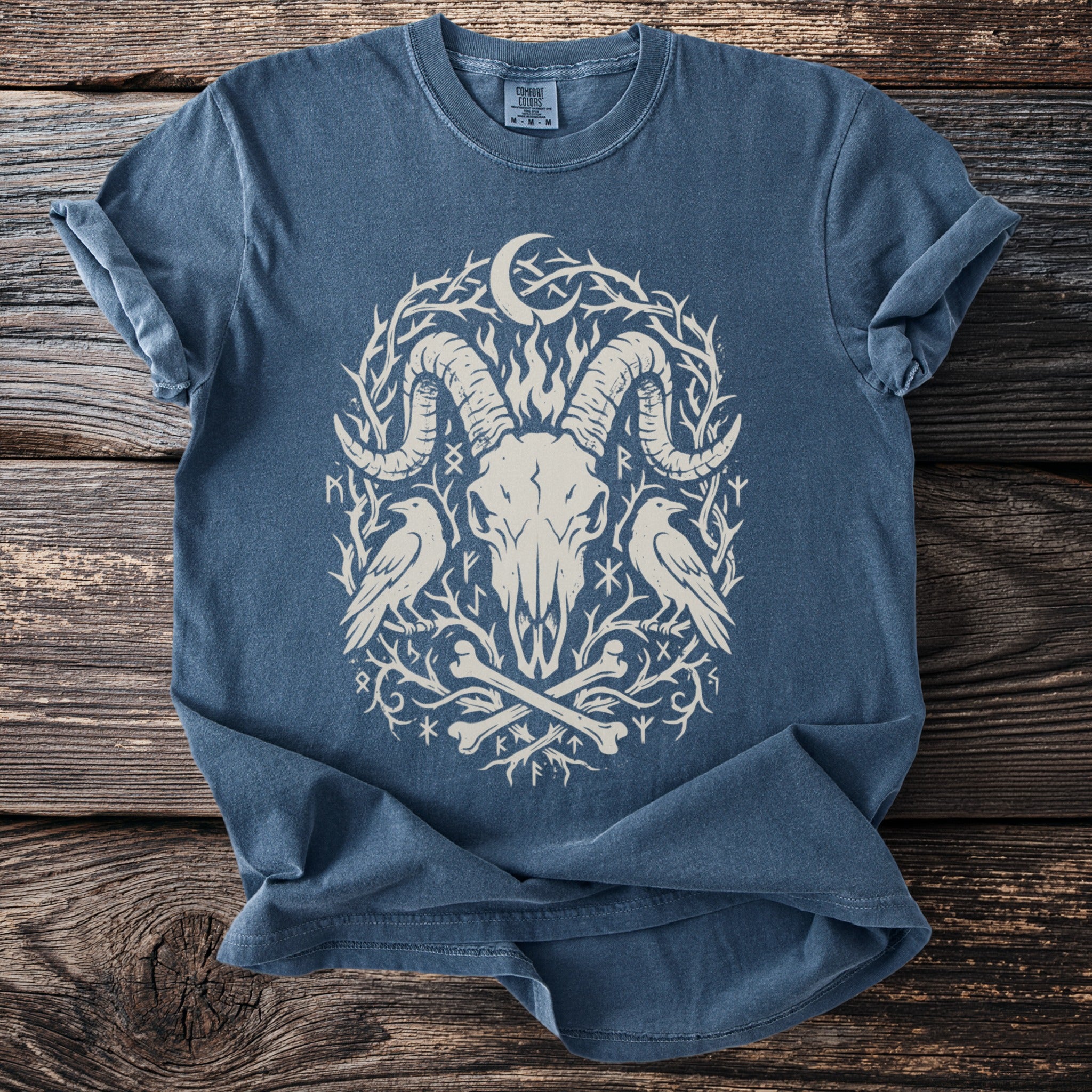 Blue t-shirt with a cream ram skull, ravens, bones, moon, and runes graphic.