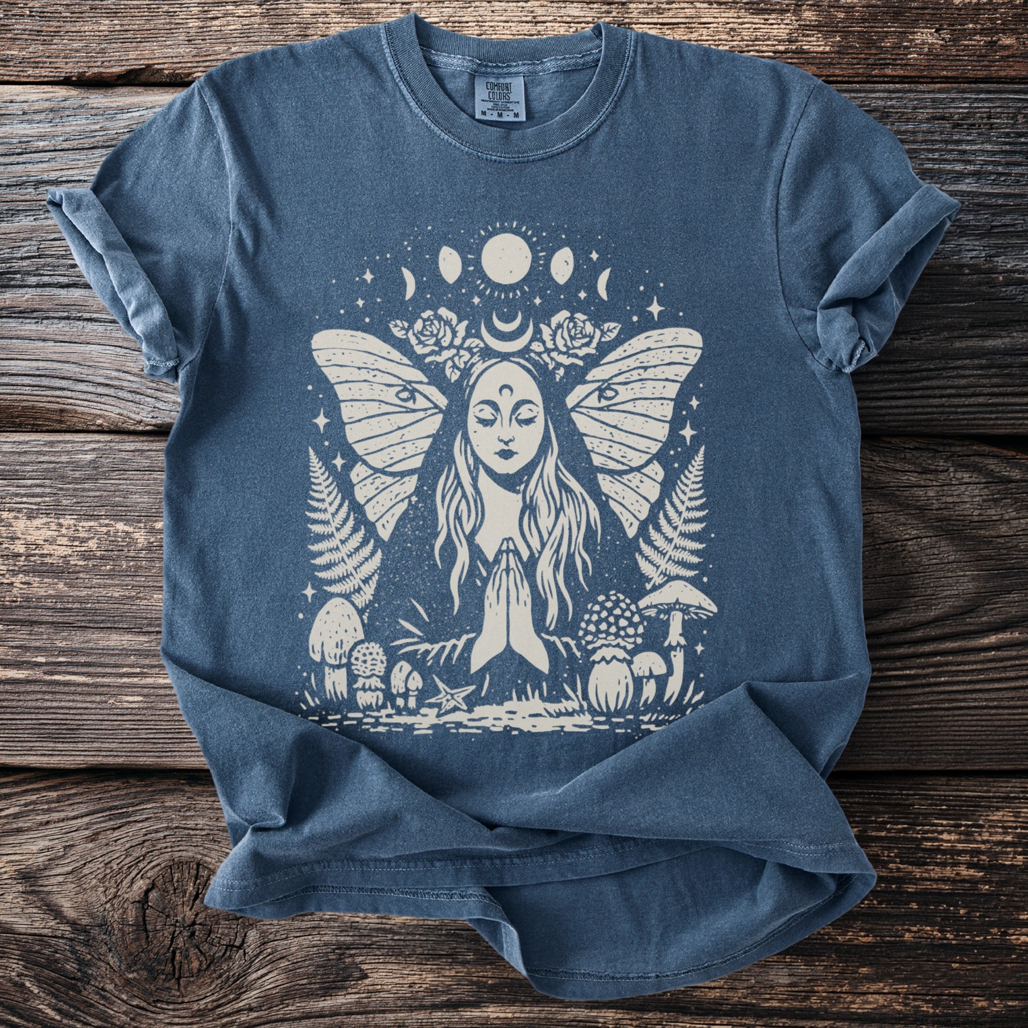 Blue t-shirt with cream print of a woman praying with moth wings and celestial elements.
