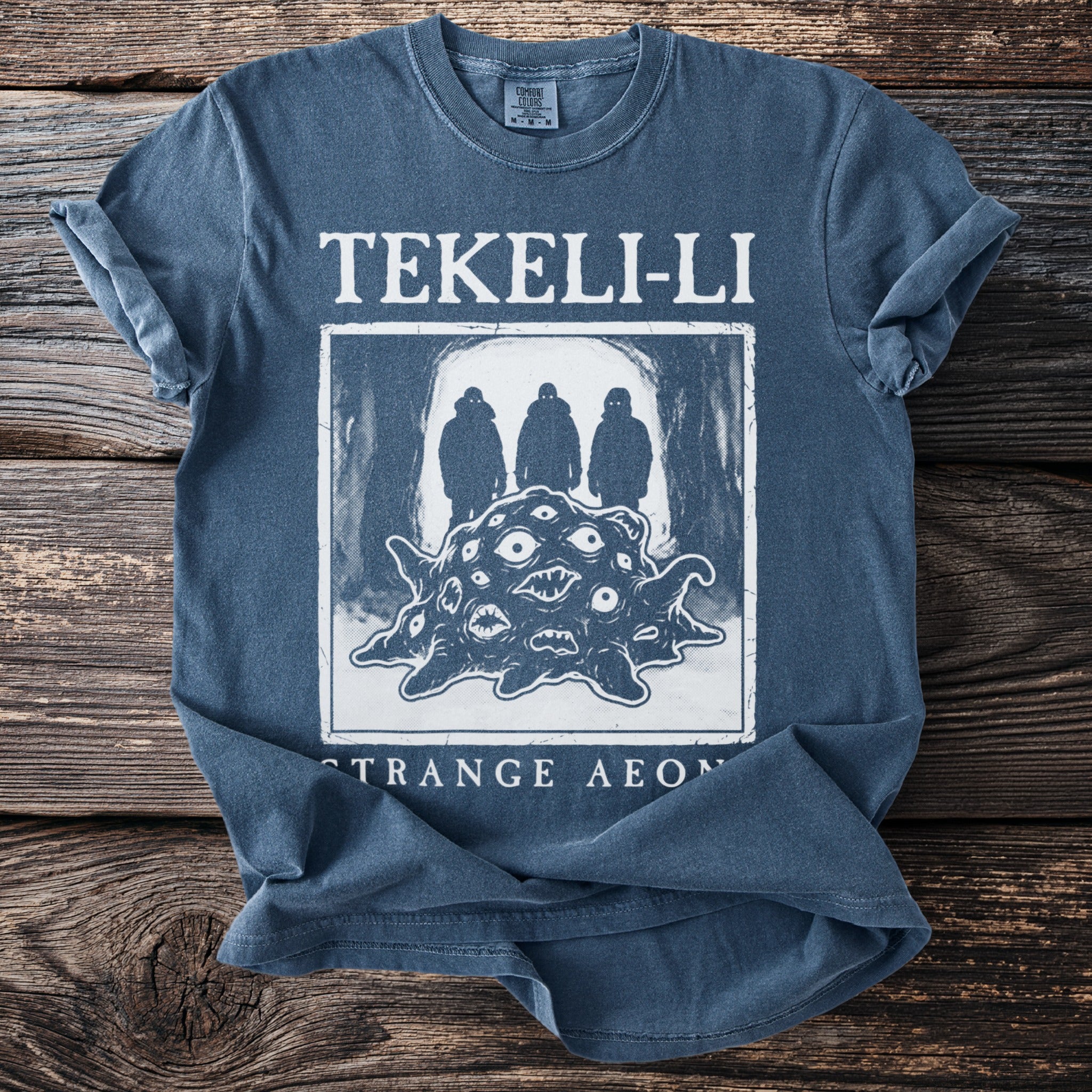Blue t-shirt with white graphic: "TEKELI-LI" above a creature with many eyes, and "STRANGE AEON" below.