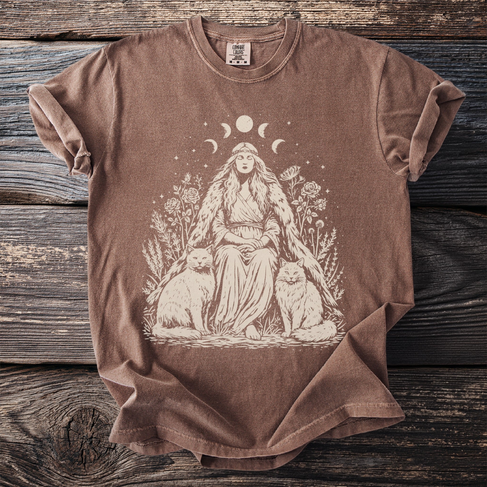Brown t-shirt with cream graphic of a woman, two cats, moon phases, and celestial accents.
