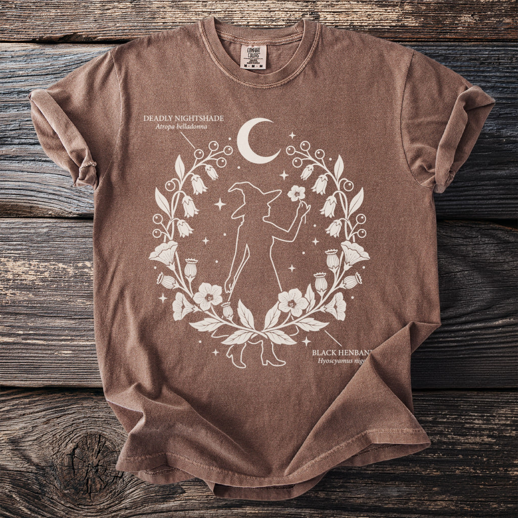 Brown t-shirt with white line art of a witch holding a flower inside a wreath.