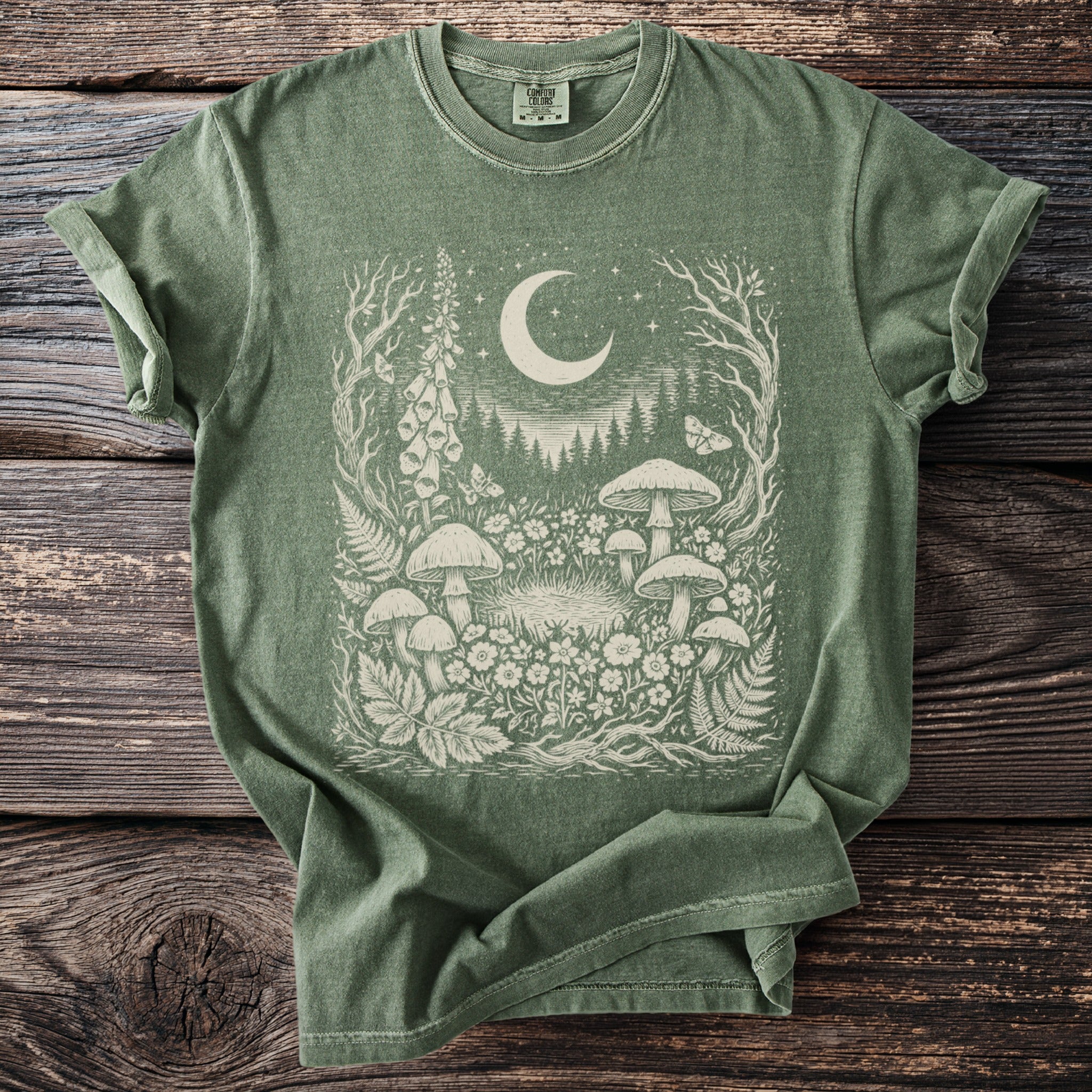 Green t-shirt with a cream graphic of mushrooms, flowers, butterflies, and a crescent moon.