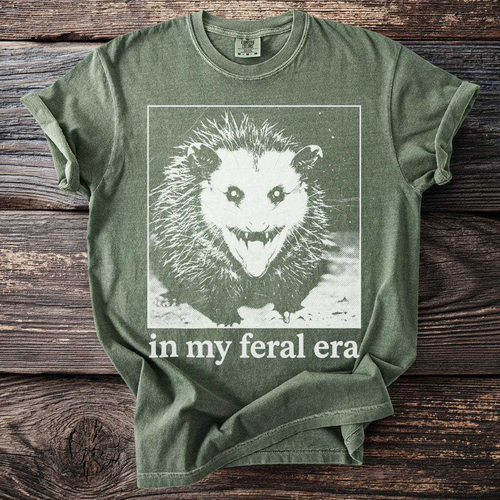 Olive green t-shirt with a distressed graphic of a snarling possum and text "in my feral era".