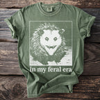 Olive green t-shirt with a distressed graphic of a snarling possum and text "in my feral era".
