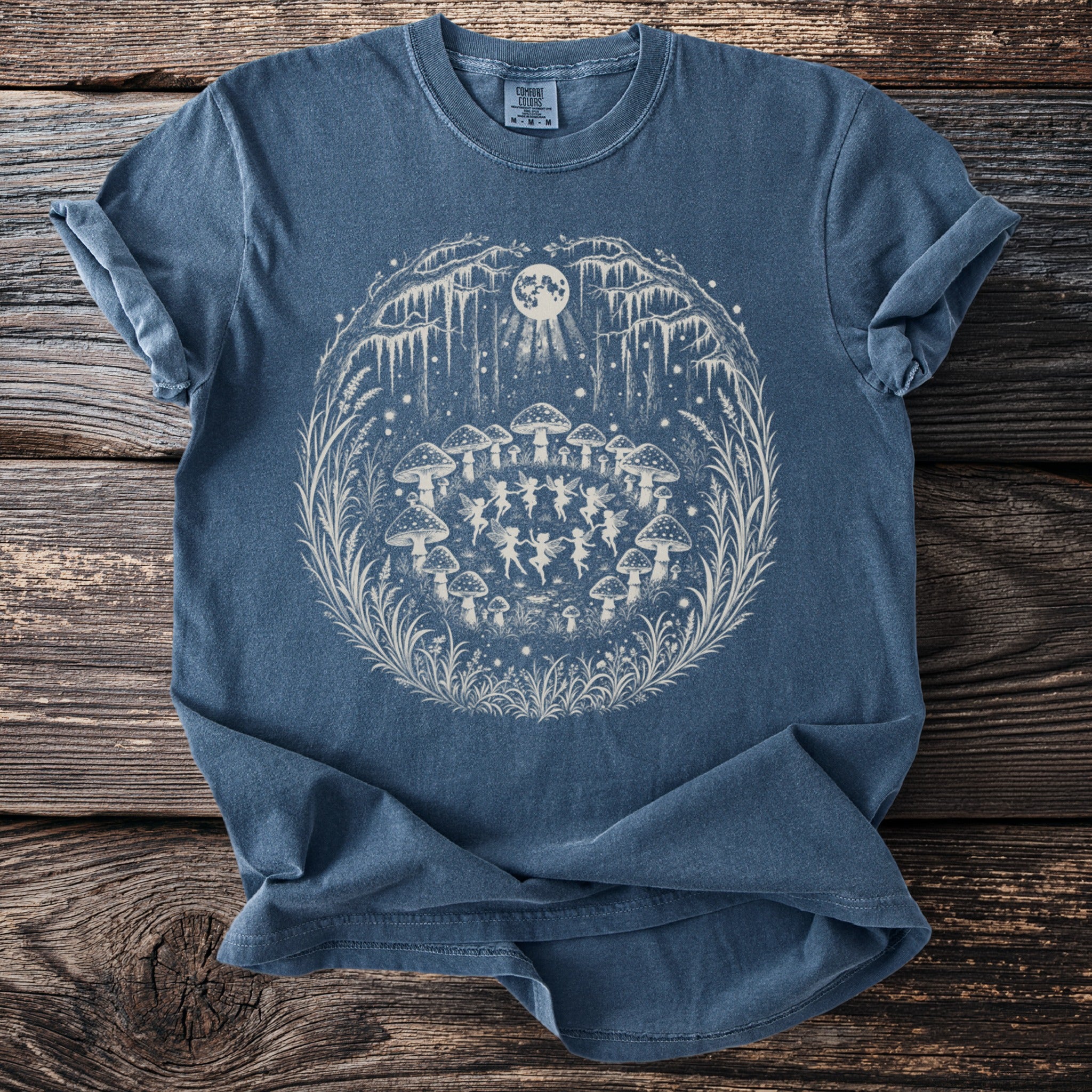 Blue t-shirt with a circular design of fairies dancing under mushrooms and a moon.