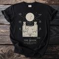 T-shirt with "The Moon" tarot card design featuring a crescent moon, towers, wolf, hound, and crayfish.