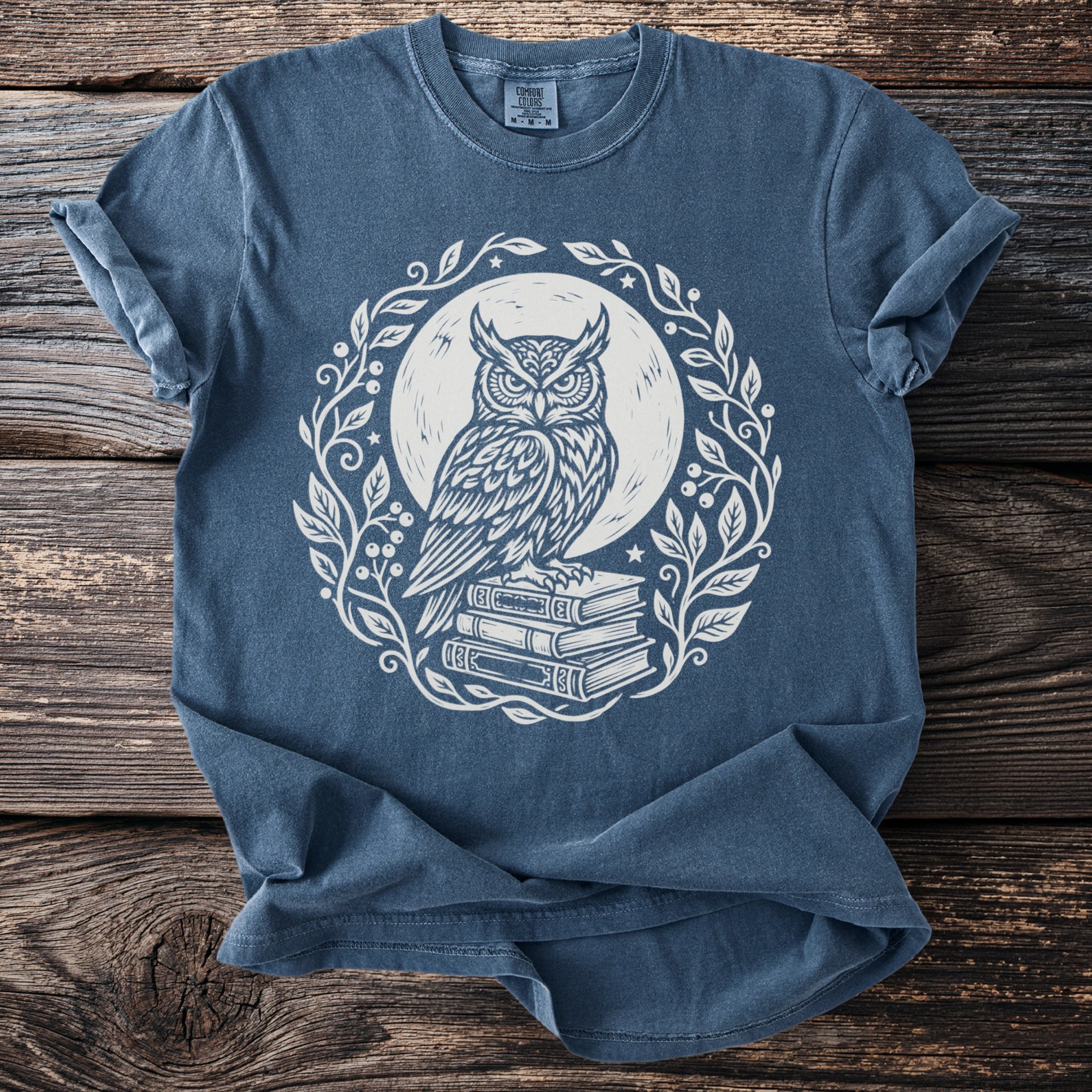 Blue t-shirt with white owl graphic sitting on books, surrounded by leaves.