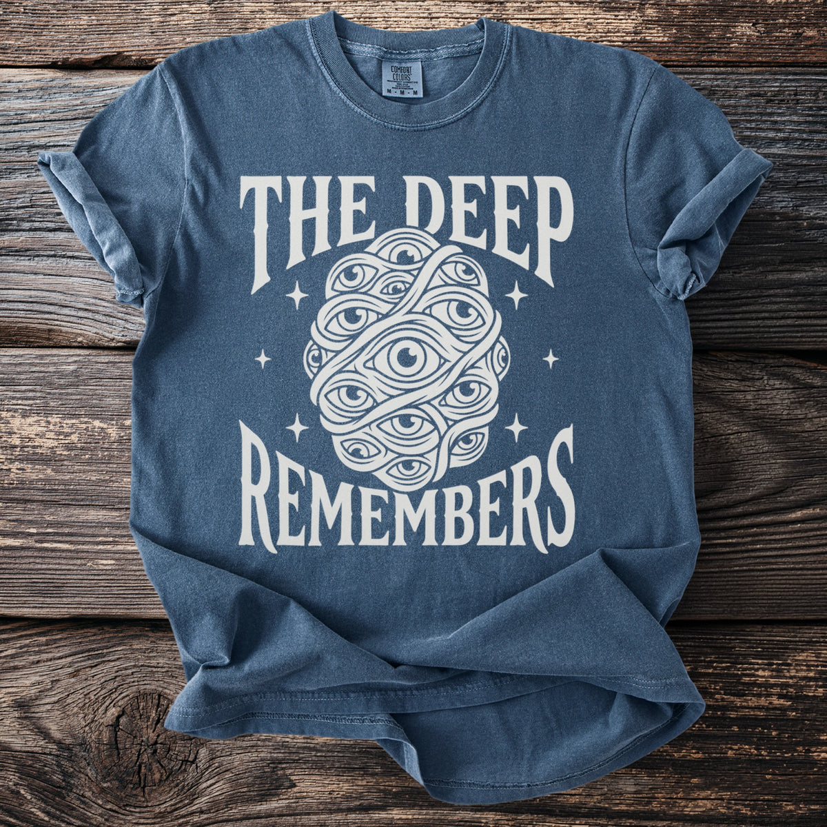Blue t-shirt with text "THE DEEP REMEMBERS" and an abstract design of eyes.