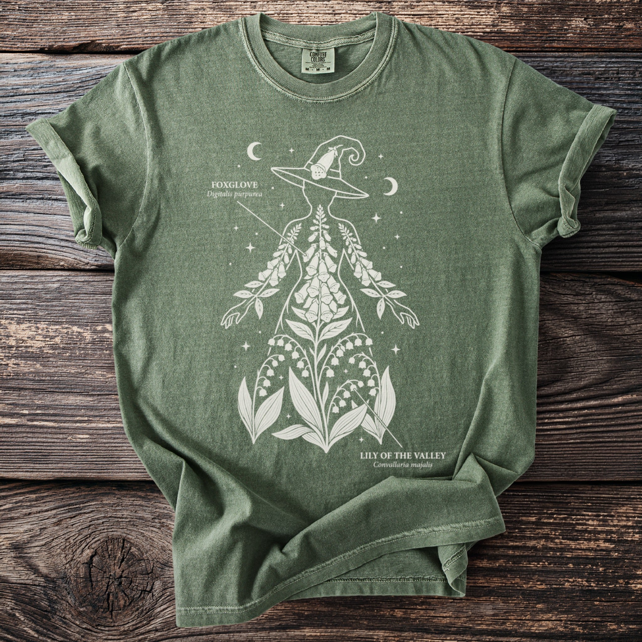 Green t-shirt with a white graphic of a witch's hat and botanical illustrations of foxglove and lily of the valley.