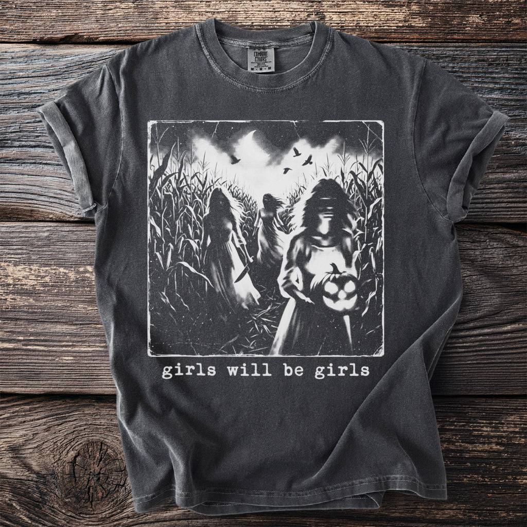 Dark t-shirt with graphic of three women in a cornfield, one holding a jack-o'-lantern, text reads "girls will be girls."