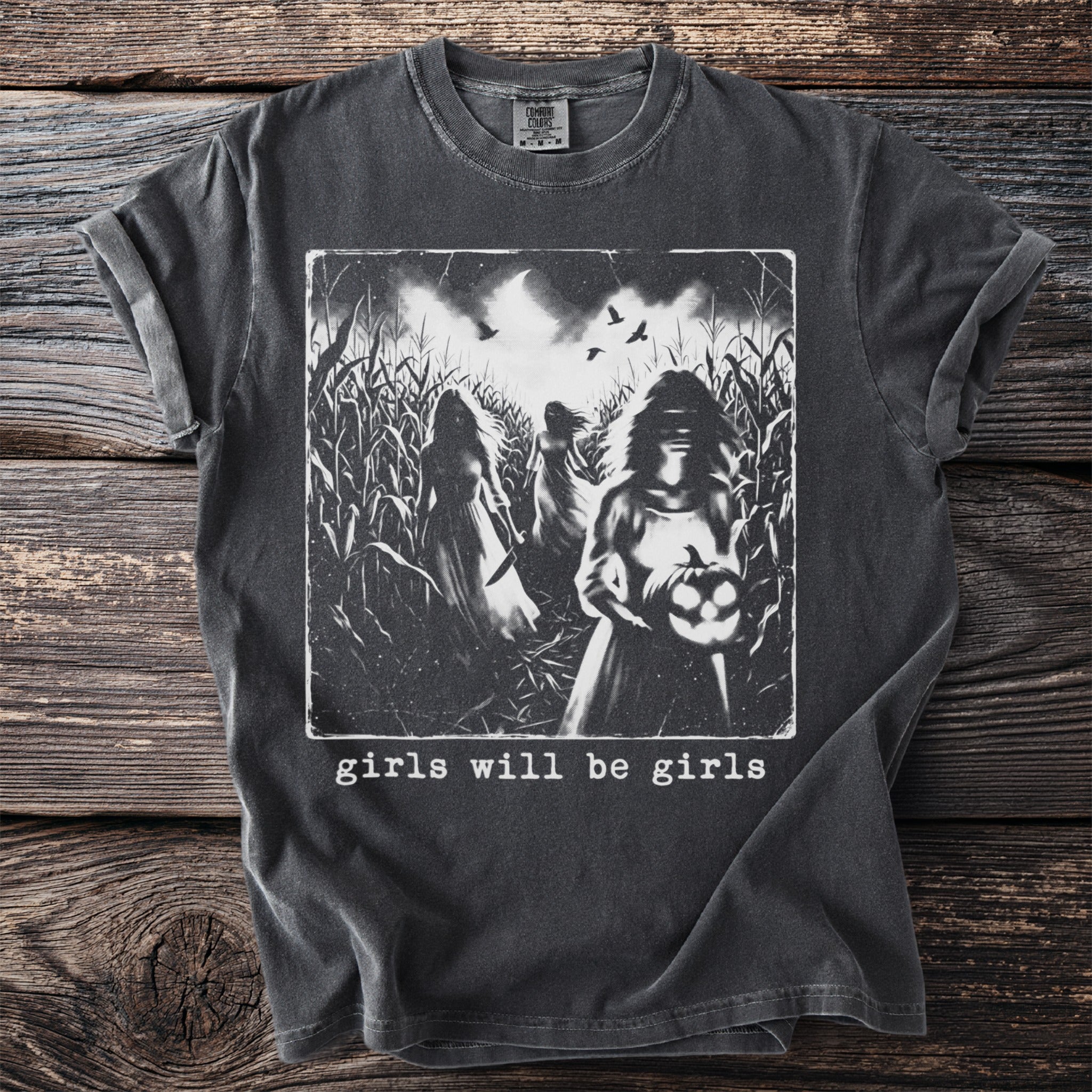 Dark t-shirt with graphic of three women in a cornfield, one holding a jack-o'-lantern, text reads "girls will be girls."